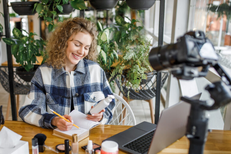 Blonde influencer with curly hair tests new products in front of her laptop and camera.