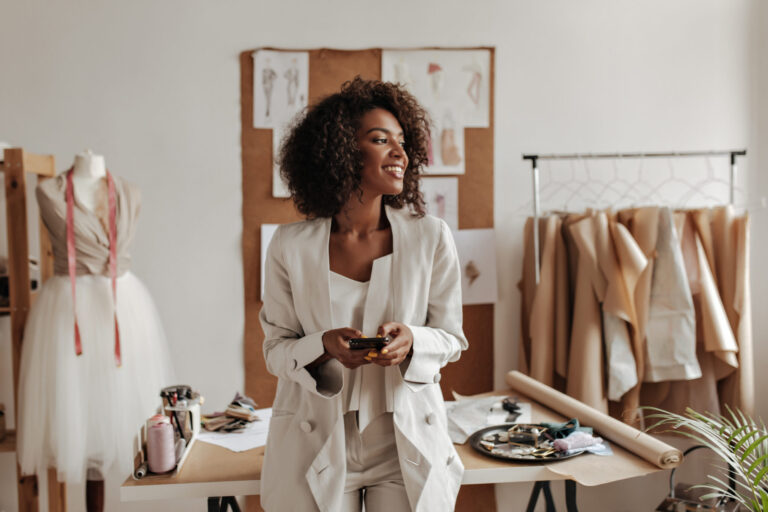 An apparel brand leader with curly brunette hair and stylish oversized white jacket smiles while holding phone and leaning on her table in her office surrounded by apparel