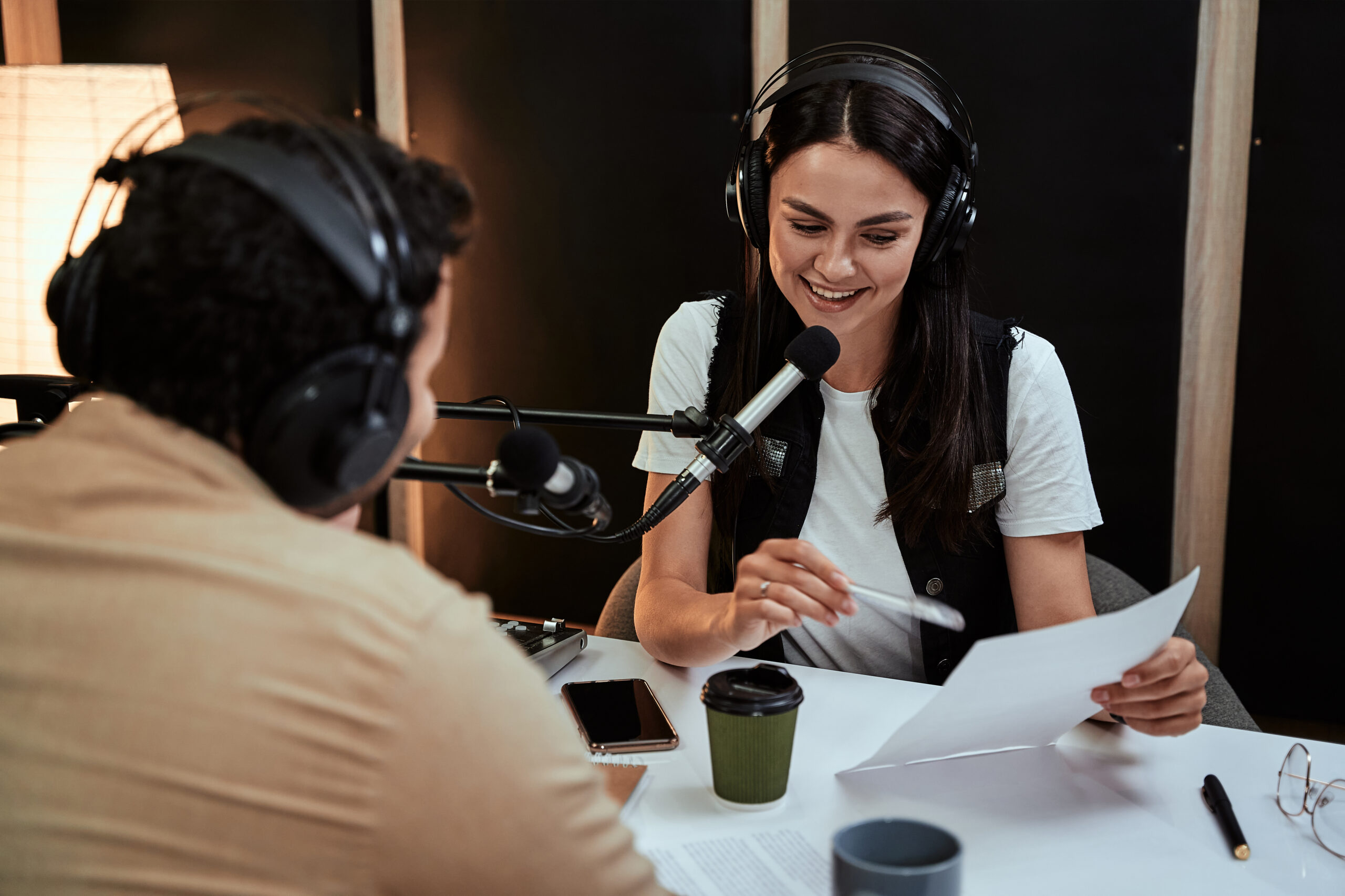 Portait of female radio host smiling, reading a script from paper while talking to male guest, presenter, moderating a live show in studio. Focus on woman. Horizontal shot
