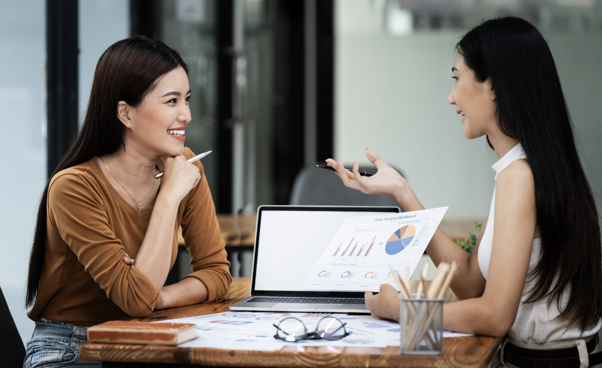 Two female business partners discuss their partnership, one holds a sheet of metrics and the other a pen.